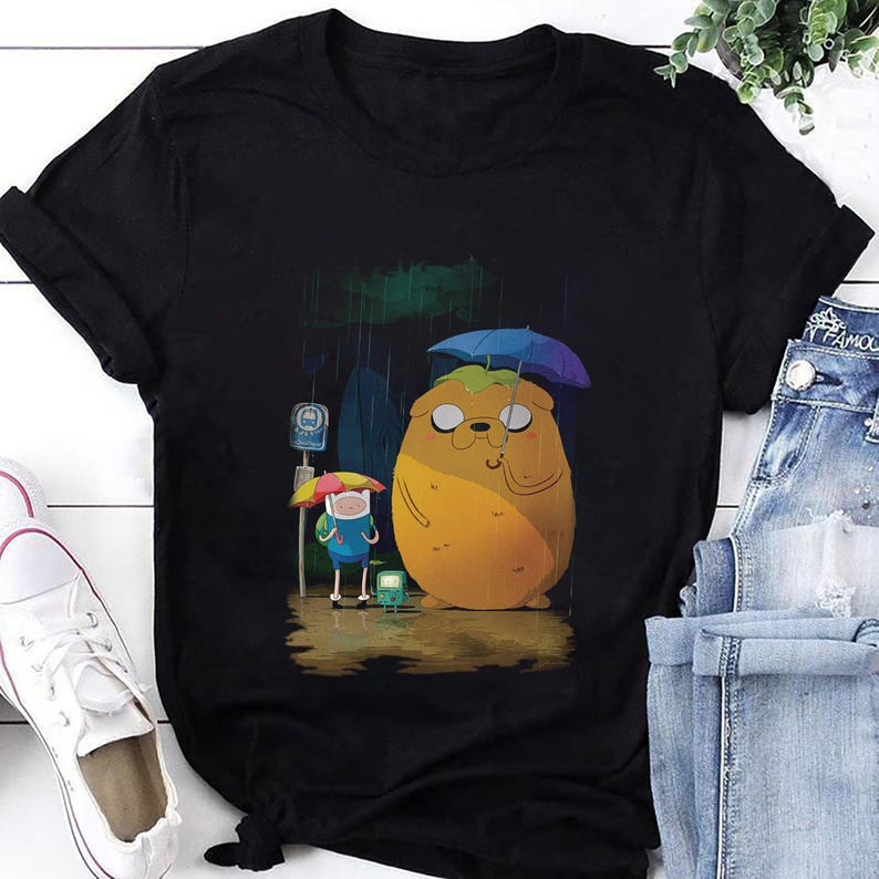 May include: Black t-shirt featuring a cartoon illustration of characters under umbrellas in the rain. The design includes a large yellow character and a smaller blue character, with a bus stop sign in the background.