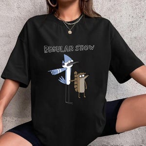 May include: Black t-shirt with the words "REGULAR SHOW" above a cartoon image of a blue jay and a raccoon. The blue jay is blue and white. The raccoon is brown and tan.