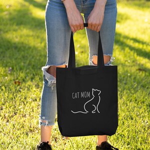 Op de afbeelding: Zwarte draagtas met de tekst "CAT MOM" en een witte kattenomtrek. De tas wordt vastgehouden door een persoon die een blauwe spijkerbroek en zwarte sneakers draagt, staand op groen gras.