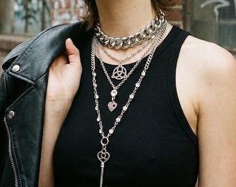 Puede incluir: Una persona lleva una camiseta sin mangas negra y varios collares plateados. Los collares incluyen una cadena gruesa, una cadena de cuentas y un collar con colgante con un dije de llave. También lleva una chaqueta de cuero negra.
