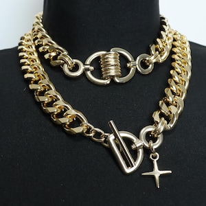 Puede incluir: Un collar de cadena de oro con una cadena grande y gruesa y una cadena más pequeña y delicada. El collar tiene un cierre grande de oro y un pequeño dije de cruz de oro.
