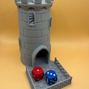May include: A grey 3D-printed dice tower, resembling a castle turret. The tower features a rectangular base with a small fence, and two dice, one red and one blue, are on the base. The tower has arched windows and a crenellated top.