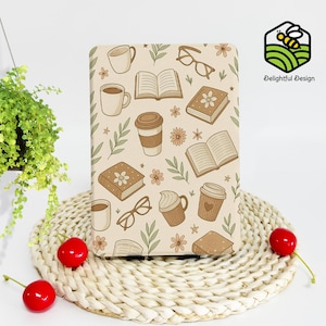 May include: A beige notebook with a repeating pattern of books, coffee cups, eyeglasses, flowers, and leaves. The notebook is rectangular and has rounded corners. The image also includes a hanging plant, a woven placemat, and two red cherries.