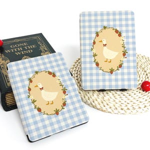 May include: Two light blue and white gingham patterned cases, each featuring a central oval design with a white duck wearing a bow tie, surrounded by strawberries and greenery. The cases are for an e-reader or tablet.