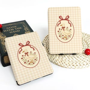 Puede incluir: Dos fundas para tabletas a cuadros beige y blancos. Cada funda presenta un diseño ovalado con una ilustración de pato, flores rojas y una cinta roja. Las fundas están sobre una estera tejida con un libro y bayas rojas en el fondo.