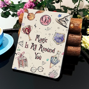 Puede incluir: Un cuaderno color crema con ilustraciones de una snitch dorada, un sombrero seleccionador y otros símbolos mágicos. El texto "Magic Is All Around You" está escrito en escritura negra. El cuaderno está rodeado de flores y una taza de café.