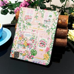 Könnte beinhalten: Ein rosa und grünes illustriertes Notizbuch mit dem Text "The Secret Garden" und einem floralen Design. Das Notizbuch ist geschlossen und liegt auf einer Holzoberfläche.