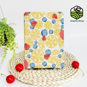 Puede incluir: Un cuaderno rectangular con fondo blanco, con un patrón de acuarela de rodajas de limón amarillas, fresas rojas y arándanos azules. El cuaderno está colocado sobre una estera tejida, con una planta colgante y cerezas en el fondo.