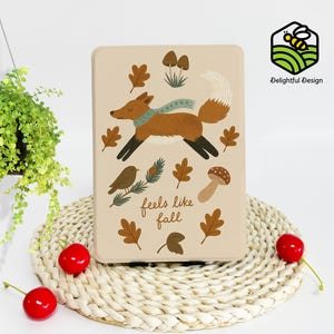 May include: A beige rectangular case featuring a running fox with a blue scarf, surrounded by autumn leaves, mushrooms, and a bird. The text "feels like fall" is displayed below the illustrations. The case has a Delightful Design logo.