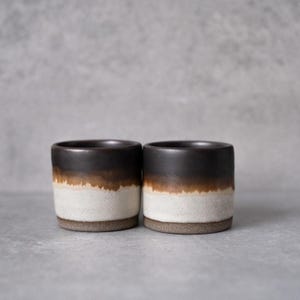 Könnte beinhalten: Zwei zylindrische Keramikbecher mit einem dunkelbraunen Rand, der in ein warmes bernsteinfarbenes Band übergeht und in einer strukturierten cremeweißen Basis endet. Die Becher haben ein glattes Finish oben und ein körniges, mattes Aussehen unten.