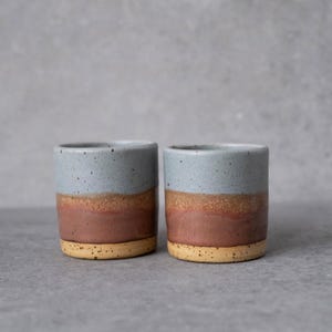 May include: Two cylindrical ceramic cups featuring a tri-colour glaze. The top section is a speckled light blue, transitioning into a warm terracotta middle band, and finishing with a speckled beige base. The cups have a smooth, straight-walled design.