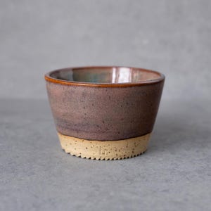 Könnte beinhalten: Eine kleine Keramikschale mit konischer Form, die eine gesprenkelte malvenfarbene und braune Glasur im oberen Bereich sowie eine texturierte, unglasierte beige Basis aufweist. Ein kleines eingestempeltes Herstellersiegel ist unten zu sehen.