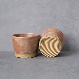 Könnte beinhalten: Zwei Keramikbecher mit gesprenkelter rosa und brauner Glasur. Ein Becher steht aufrecht, während der andere gekippt ist und einen Boden mit texturiertem, geripptem Muster zeigt. Ein kleines, quadratisches Herstellersiegel ist unten eingeprägt.