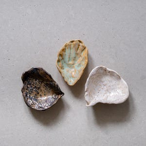 Può includere: Tre piccole ciotole in ceramica di diversi colori e texture. Le ciotole hanno la forma di conchiglie e hanno un aspetto rustico e fatto a mano.