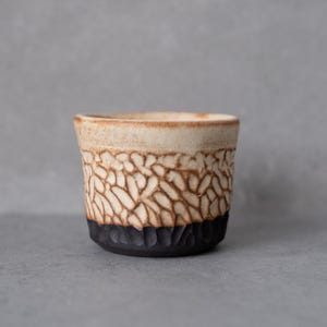 Könnte beinhalten: Ein Keramikbecher mit zweifarbigem Design, bestehend aus einem glatten, hellbeigen oberen Teil und einem dunklen, strukturierten schwarzen Boden. Der Mittelteil zeigt ein geschnitztes, sich wiederholendes Muster aus länglichen, organischen Formen mit braunen Akzenten.