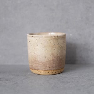 Könnte beinhalten: Ein zylindrischer Keramikbecher mit einer gesprenkelten cremefarbenen Glasur im oberen Bereich, die zum Boden hin in einen warmen, erdigen Braunton übergeht. Das Gefäß hat ein glattes, geradwandiges Profil mit flachem Boden und ohne Henkel.