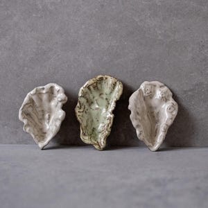 Può includere: Un set di tre tazze in ceramica con finitura opaca e macchiettata. Le tazze sono nei toni del crema, grigio chiaro e grigio scuro. Ogni tazza ha un design semplice e minimalista.
