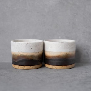 May include: Two cylindrical ceramic cups featuring a speckled white upper section that transitions into a smooth gradient of tan, brown, and dark charcoal at the base. The cups have a straight-sided design with a flat bottom and a speckled texture.