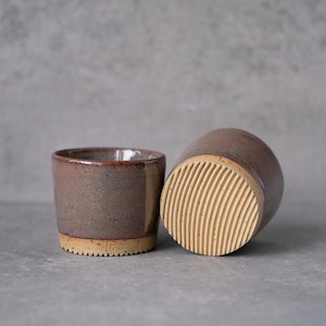 May include: Two ceramic cups with a speckled mauve and brown glaze. One cup stands upright, showing a smooth interior and a textured, ribbed base. The second cup is tilted on its side, revealing a light beige, deeply grooved, circular bottom.