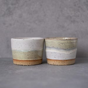 May include: Two cylindrical ceramic cups featuring a speckled white and sage green glaze with a smooth transition between colours. The base of each cup is unglazed, showing a natural tan clay texture with a small stamped maker mark near the bottom.