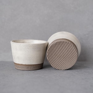 May include: Two ceramic cups with a speckled off-white glaze on the upper portion and a dark, textured, ribbed base. One cup is positioned upright, while the other is tilted on its side to reveal the circular, grooved pattern on the bottom.