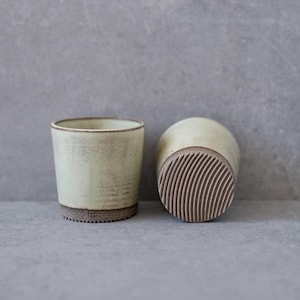 Può includere: Due tazze in ceramica bianco sporco con base strutturata e scanalata e bordo marrone scuro. Una tazza è in posizione verticale, l'altra è di lato, rivelando il fondo a motivi. Le tazze hanno un design minimalista.