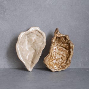 Puede incluir: Dos cuencos de cerámica con forma de concha de ostra. El cuenco de la izquierda tiene un acabado marmolado liso y brillante en crema y marrón claro. El de la derecha presenta un esmalte texturizado en marrón oscuro con hendiduras.