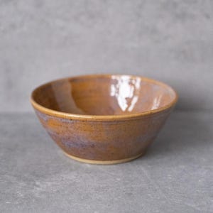 Könnte beinhalten: Eine runde Keramikschale mit glänzender Oberfläche, die eine warme bernsteinfarbene und goldbraune Glasur mit subtilen violetten Untertönen nahe der Basis aufweist. Die Oberfläche hat eine gesprenkelte Textur, einen glatten Rand und eine konische Form.