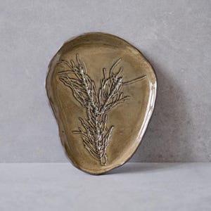 Pode incluir: Uma travessa de cerâmica artesanal com uma forma orgânica única. A travessa tem uma cor marrom quente e terrosa com um acabamento brilhante. Um design botânico em relevo detalhado de uma planta está centrado na travessa.