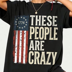 Może przedstawiać: Czarny t-shirt z postarzanym wzorem amerykańskiej flagi, rokiem "1776" i napisem "THESE PEOPLE ARE CRAZY" w stylu vintage. Koszulka ma krótkie rękawy i luźny krój, idealna do noszenia na co dzień.