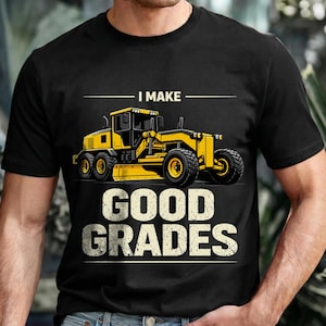 Puede incluir: Camiseta negra con un diseño de motoniveladora amarilla y el texto "I MAKE GOOD GRADES". La motoniveladora, un vehículo de construcción, se representa con neumáticos grandes y una cuchilla. La camiseta es de cuello redondo.