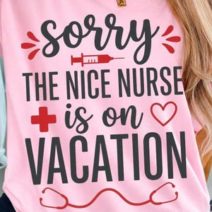 以下が含まれることがあります： 「Sorry The Nice Nurse is on Vacation」と書かれたピンク色のTシャツ。 黒い文字に、注射器、十字、ハート、聴診器のデザインが赤色でアクセントとして加えられています。