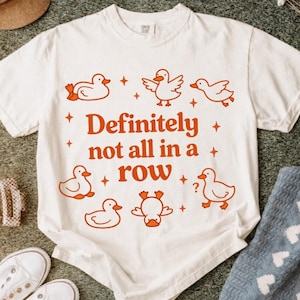 May include: White t-shirt with orange duck illustrations and the phrase "Definitely not all in a row." The design includes ducks in various poses and small star accents. A casual, fun graphic tee.