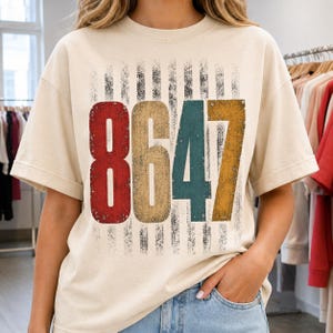 May include: Cream-colored t-shirt featuring the numbers "8647" in a distressed, vintage style. The numbers are in red, tan, teal, and gold, set against a backdrop of vertical black stripes. The shirt has short sleeves.