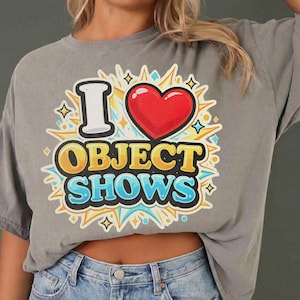 Könnte beinhalten: Graues T-Shirt mit dem Text "I ❤️ OBJECT SHOWS" in einem farbenfrohen Retro-Design. Die Grafik enthält ein rotes Herz, gelbe und blaue Akzente und eine weiße Umrandung. Das Shirt besteht aus einem weichen Material.