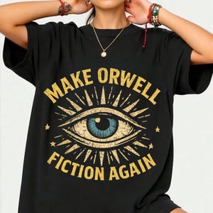 Puede incluir: Camiseta negra con un gráfico dorado desgastado que dice "MAKE ORWELL FICTION AGAIN". El gráfico presenta un ojo con un iris azul y líneas radiantes. Dos pequeñas estrellas flanquean el diseño del ojo.