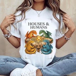 May include: White t-shirt with the text "HOUSES & HUMANS" and a colourful illustration. The image shows four dragons, orange, green, yellow, and blue, gathered around a table with a central fire pit.