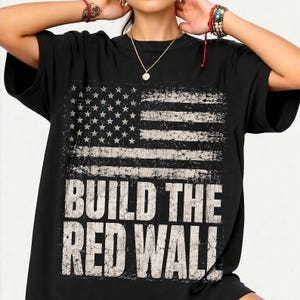 Könnte beinhalten: Schwarzes T-Shirt mit einem verwitterten amerikanischen Flaggen-Design in Weiß und Grau. Der Text "BUILD THE RED WALL" ist darunter gedruckt. Das T-Shirt hat einen Rundhalsausschnitt und kurze Ärmel.