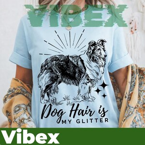Könnte beinhalten: Hellblaues T-Shirt mit einer schwarz-weißen Illustration eines Hundes und dem Text "Dog Hair is My Glitter". Das Design beinhaltet einen Sonnenaufgang und Sterne, ideal für Hundeliebhaber.