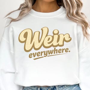 May include: White sweatshirt with the text "Weir everywhere" in a retro font. The word "Weir" is in a large, tan, rounded font, and "everywhere" is in a smaller, sans-serif font below it. The sweatshirt is a casual style.