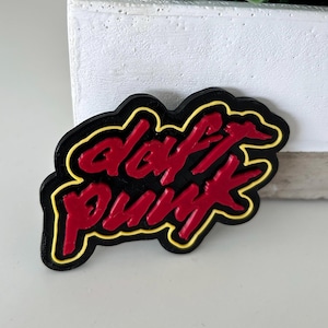 Könnte beinhalten: Ein schwarzer und roter Daft Punk Logo-Pin mit gelber Umrandung. Der Pin zeigt den Schriftzug "Daft Punk" in einer markanten Schriftart. Der Pin ist wahrscheinlich aus Kunststoff oder Metall, für Kleidung oder Accessoires.