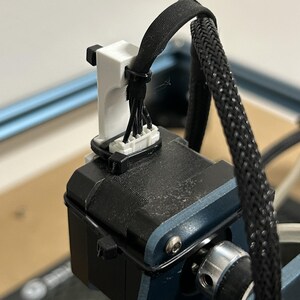 May include: A close-up of a 3D printed white cable management clip attached to a black stepper motor. The clip is holding a bundle of black wires.