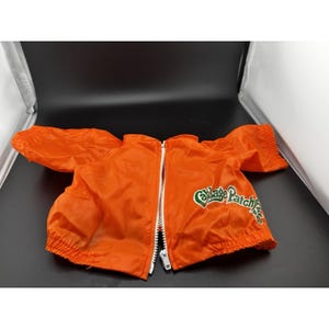 May include: Bright orange nylon jacket with a white zipper and elastic cuffs. The jacket has the words "Cabbage Patch Kids" in green script on the right side. The jacket is designed for a doll.