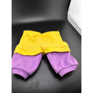 Può includere: Un paio di pantaloni della tuta per bambini, con un design a blocchi di colore. La parte superiore è gialla, mentre le gambe inferiori sono viola. I pantaloni sono realizzati in un materiale morbido, forse in pile.