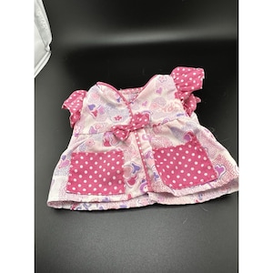 Könnte beinhalten: Ein rosa-weiß gemustertes Puppenkleid mit Schleife und zwei gepunkteten Taschen. Das Kleid hat ein Herz- und Blumenmuster mit rosa Tupfen auf den Taschen und Ärmeln.