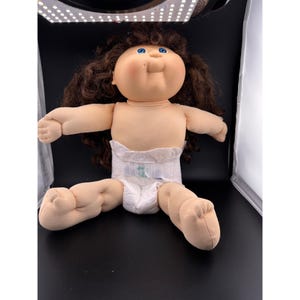 May include: A vintage Cabbage Patch Kids doll with brown hair and blue eyes. The doll is wearing a white diaper and has a soft, plush body. The doll is sitting with its arms and legs outstretched.