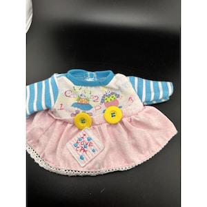 Könnte beinhalten: Ein Puppenkleid mit einem weißen Oberteil und blau gestreiften Ärmeln, kombiniert mit einem rosa Rock. Das Oberteil ist mit einem Cartoon-Design und den Zahlen 1 und 2 sowie gelben Knöpfen verziert. Ein kleiner Aufnäher mit dem Text "I LOVE CPK" ist auf den Rock genäht.