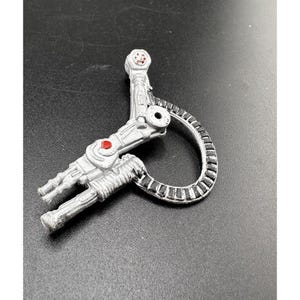 May include: A silver-colored, mechanical-looking keychain with a circular, segmented design and red accents. The keychain has a detailed, industrial aesthetic, with a unique, stylized shape.