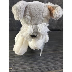 May include: A plush Schnauzer dog toy with gray and white fur. The toy has floppy ears, a long white beard, and a black nose. A clear tag is attached to the toy.