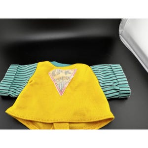 May include: A yellow doll shirt with teal and black striped sleeves. The shirt features a pink, white, and grey triangle patch on the front with the letters "CFK" and other text. The shirt is made of a soft material.